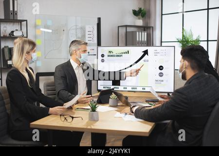 Mature bearded chief in face mask, having a meeting together with his focused international colleagues, and showing business strategy on interactive digital screen with financial marketing charts. Stock Photo