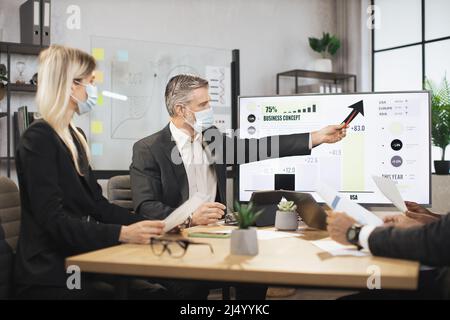 Mature bearded chief in face mask, having a meeting together with his focused international colleagues, and showing business strategy on interactive digital screen with financial marketing charts. Stock Photo