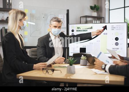 Mature bearded chief in face mask, having a meeting together with his focused international colleagues, and showing business strategy on interactive digital screen with financial marketing charts. Stock Photo