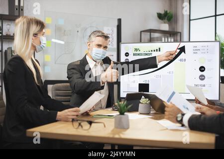 Mature bearded chief in face mask, having a meeting together with his focused international colleagues, and showing business strategy on interactive digital screen with financial marketing charts. Stock Photo