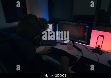 Young blond guy playing video games on a computer with headphones and using a backlit coloured keyboard. Stock Photo