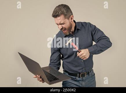 Furious man break laptop hitting notebook with hammer grey background, computer rage Stock Photo