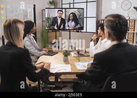 Team of diverse four multiethnic businesspeople having video meeting in boardroom at office indoor looking at African business lady and Caucasian man, using smart video technology to communicat Stock Photo