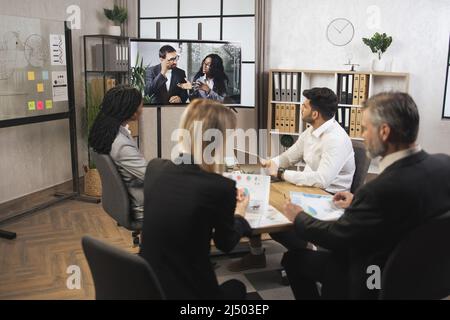 Team of diverse four multiethnic businesspeople having video meeting in boardroom at office indoor looking at African business lady and Caucasian man, using smart video technology to communicat Stock Photo