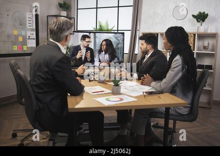 Focused multiracial business people in conference room, looking at a screen, having video conference with another team, African lady and Caucasian man, using smart video technology to communicate. Stock Photo