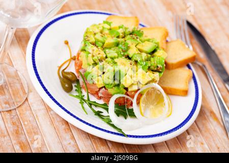 Salmon tartare with avocado served with toasts, greens, lemon and capers Stock Photo