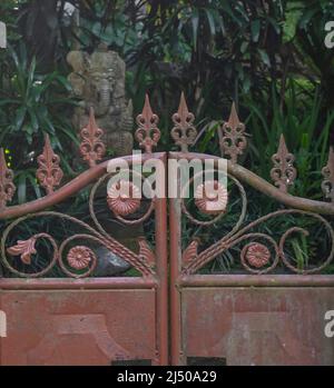 Old red iron gate with flower emblems and blurred background with Balinese ganesha concrete statue among green plants Stock Photo