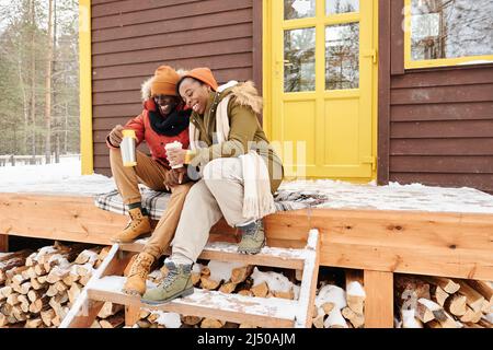 Young cheerful and affectionate husband and wife with drinks sitting on wooden porch of their country house and enjoying winter weekend Stock Photo