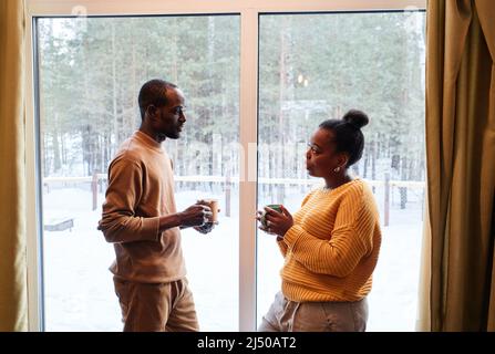 Side view of young African American couple in casualwear having tea while standing in front of one another by window of country house Stock Photo