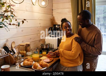 Happy young black woman slicing tomatoes and looking at husband standing behind and keeping his hands on her shoulders Stock Photo