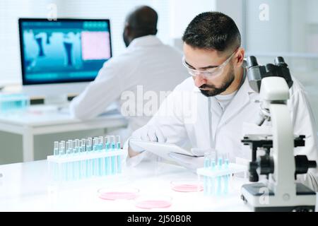 Contemporary young male scientist in workwear using digital tablet while searching for online data about new biotechnologies Stock Photo