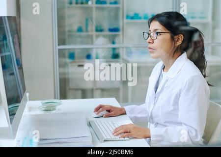 Young pretty female scientist in whitecoat and eyeglasses using computer for studying new virus while sitting by workplace in laboratory Stock Photo