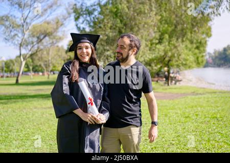 Young girl recently graduated, dressed in cap and gown, with her degree in her hands, celebrating with her family on the university campus. Very happy Stock Photo