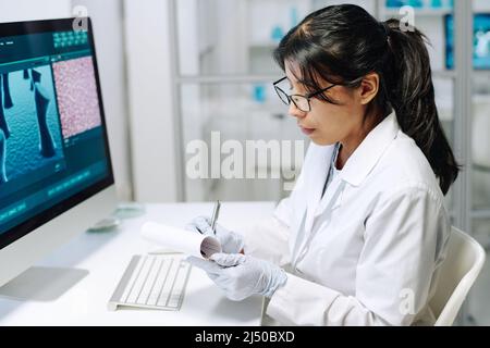 Young serious Hispanic female scientist making notes in her notepad while sitting by workplace in front of computer during research Stock Photo