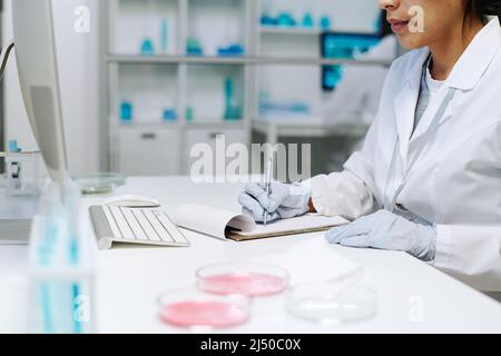 Gloved hands of young female scientist in whitecoat making notes in clinical document while sitting by workplace in laboratory