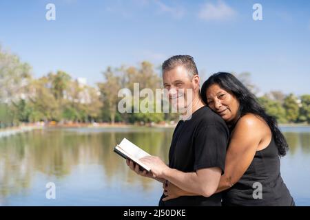 Mature multi ethnic couple embracing by a lake reading a book, smiling looking at camera. Copy space Stock Photo