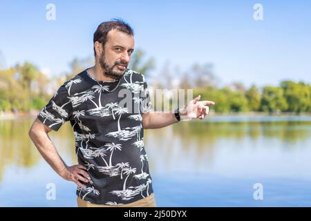 Bearded mature man in a lake looking at the camera with one arm akimbo and the other pointing to the side. Copy space Stock Photo