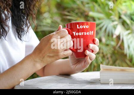 Woman holding a red cup with text Love Yourself, and a book on table. Outdoor with palm tree background. Stock Photo