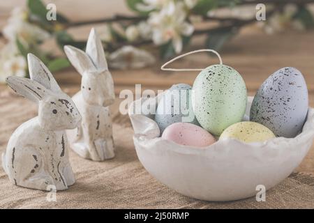 Decorative Easter eggs in pastel colors in a white handmade bowl and two wooden Easter bunnies on a wooden table, Easter decoration Stock Photo
