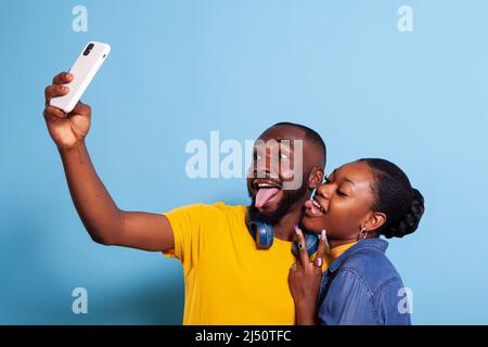 Goofy couple sticking tongue out and taking pictures on smartphone, having fun with photography. Silly man and woman fooling around with selfies, making memories on mobile phone. Stock Photo