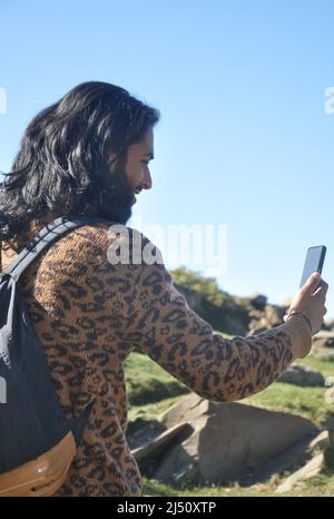 Rear view of a good looking long haired young man taking selfie or having video call with his smartphone in the mountain Stock Photo