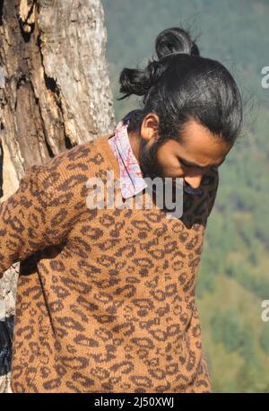 Closeup of a good looking Indian young men with hair bun looking down while leaning against tree Stock Photo
