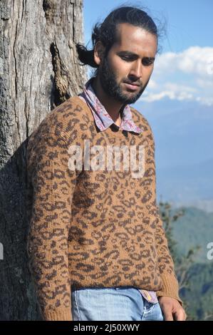 A south asian young man looking down while leaning against tree in the nature Stock Photo