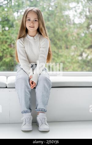 full length view of girl in jeans sitting on windowsill and looking at camera Stock Photo