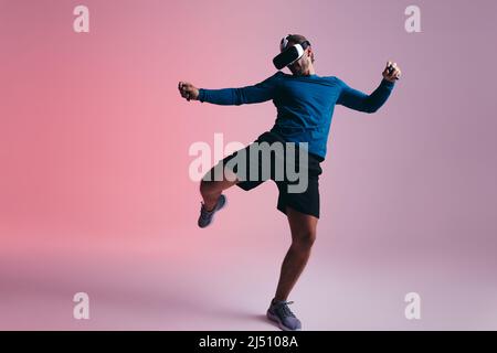 sporty young man kicking a virtual ball while wearing virtual reality goggles. Young man playing football in virtual reality. Active young man explori Stock Photo