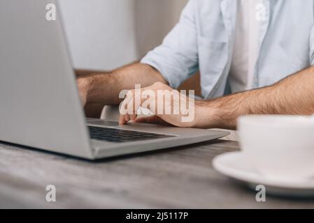 Hands of a modern young programmer or office manager over a laptop keyboard while programming. A guy in a blue shirt and white T-shirt. Cup of coffee on the table. Freelance Stock Photo