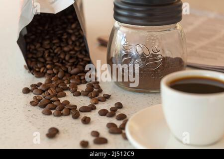 Coffee beans spill from their packet on a benchtop with a cup of coffee and ground coffee nearby. Stock Photo