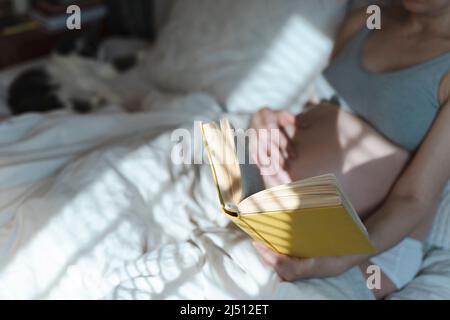 A pregnant woman reads a book and strokes her belly with a baby before childbirth while relaxing in a sunny bright bedroom lying on a bed. Selected focus. Women's health, pregnancy, conception, childbirth concept. High quality photo Stock Photo