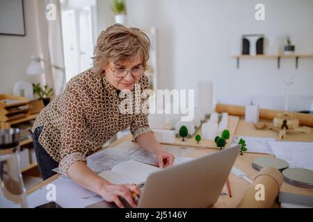 Senior woman architect with model of houses working on laptop in office. Stock Photo