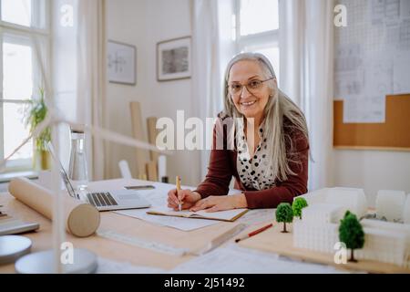 Senior woman architect with model of houses in office writing notes. Stock Photo