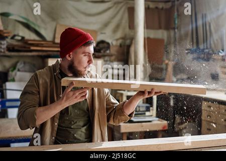 Modern male carpenter wearing red knit cap holding wood plank blowing dust from it in joinery workshop Stock Photo