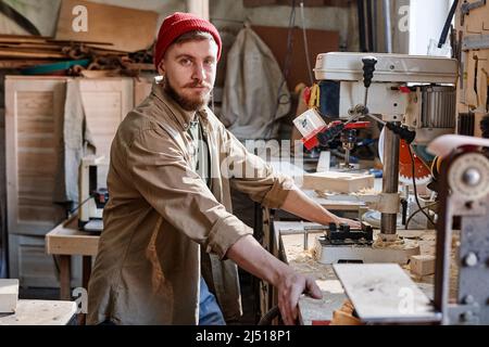 Portrait of young Caucasian male carpenter with beard on face working with drill press looking at camera Stock Photo