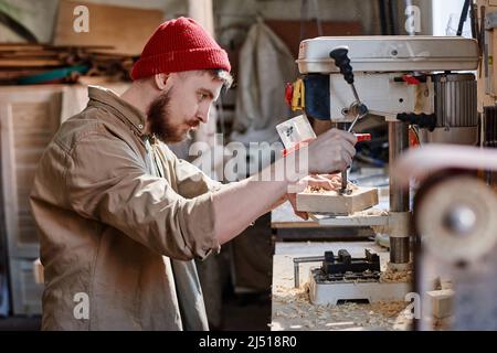 Side view of modern young male carpenter using drill press to make holes in wooden object in woodworking workshop Stock Photo
