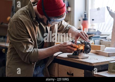 Professional young male carpenter wearing safety glasses working in joinery workshop sharpening knife using grinding jig Stock Photo