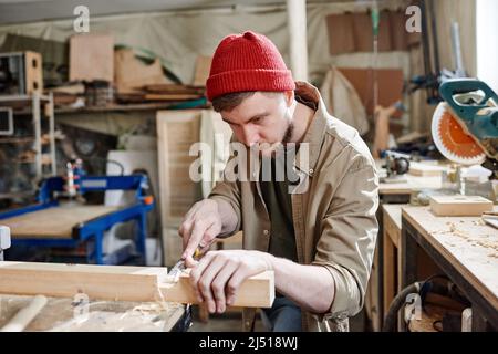Bearded Caucasian carpenter wearing red knit cap cutting mortise in wood plank using chisel Stock Photo