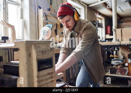 Young male carpenter wearing noise-cancelling headphones using thickness planer machine in woodworking workshop Stock Photo