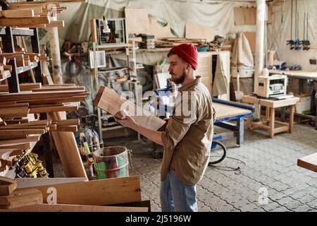 Young Caucasian man working in carpentry workshop choosing and taking out wood plank from shelf Stock Photo