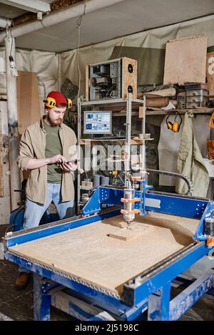 Vertical long shot of young Caucasian man using tablet while operating CNC milling machine in woodworking workshop Stock Photo