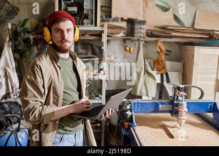 Horizontal medium portrait of young Caucasian man working in carpentry workshop holding laptop looking at camera Stock Photo