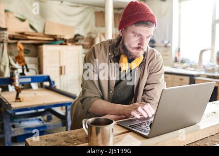 Young bearded carpenter with hearing protection headphones over his neck surfing Internet on laptop during coffee break Stock Photo