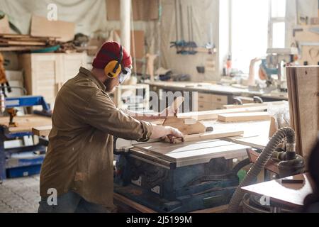Unrecognizable carpenter wearing safety glasses and earmuffs cutting wooden board with table saw Stock Photo