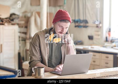 Caucasian male carpenter talking on phone while texting something on laptop during workday in joinery workshop Stock Photo