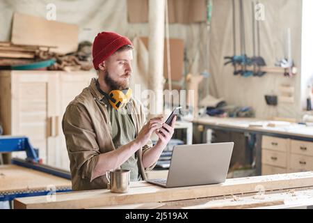 Modern woodworker standing at workbench with laptop and cup of tea on it texting something on smartphone Stock Photo