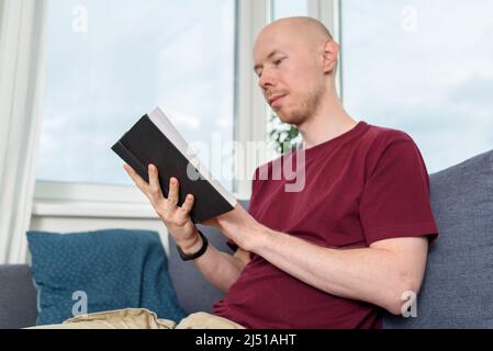 Bald hairless man reading book sitting on couch Stock Photo
