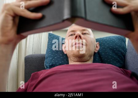 Bald hairless man reading book lying on couch Stock Photo