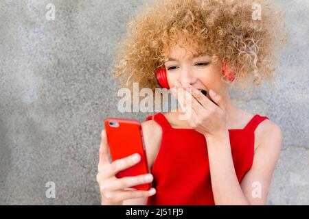 young latin woman with curly blonde hair using phone with headphones with a surprised expression wearing a red dress Stock Photo